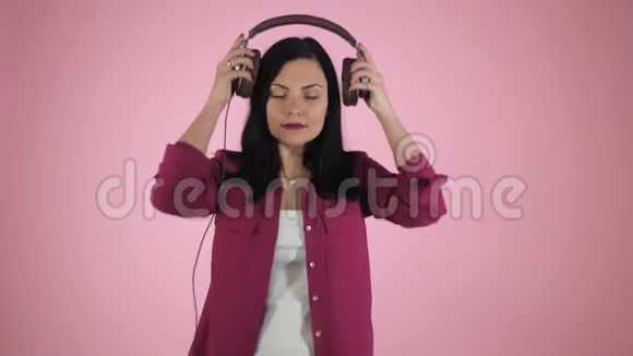 美丽的年轻黑发女人戴着耳机听音乐在彩色背景上唱歌粉红色的快乐女孩视频的预览图