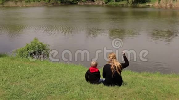 妈妈和儿子正坐在春天河岸上的绿草上视频的预览图