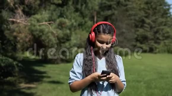 一个长发迷人的女孩走过公园用耳机听音乐慢吞吞的视频的预览图