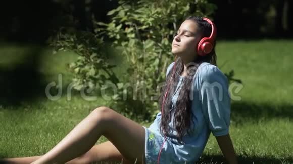 一个年轻的女孩留着长长的黑发坐在公园的草地上用平板电脑听着耳机里的音乐玩得很开心视频的预览图