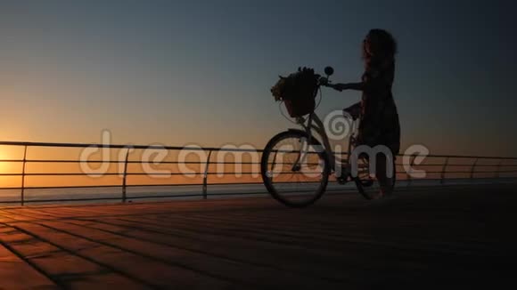 穿着老式自行车和花束的年轻女子在海边木堤上行走的剪影视频的预览图