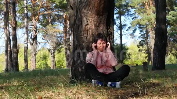 年轻女子坐在公园的一棵树旁通过耳机听音乐音乐户外娱乐视频的预览图