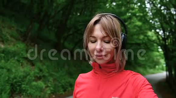 一位戴着耳机的漂亮金发女孩在慢跑和看她的健康状况之前站在森林里特写镜头视频的预览图