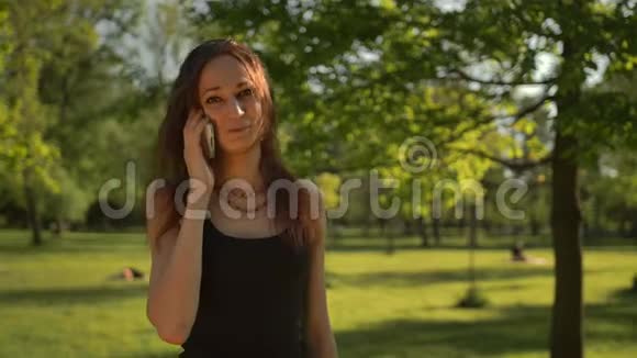 微笑的年轻女子站在公园户外通过电话交谈视频的预览图