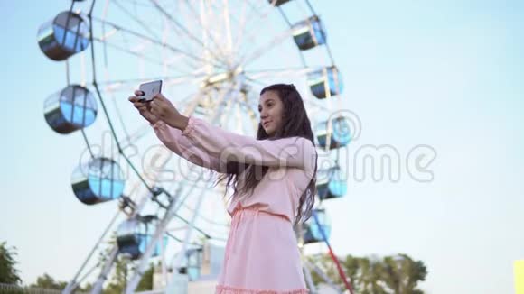 一个穿着粉红色长裙的长发女孩在摩天轮附近站着用智能手机自拍4K视频的预览图