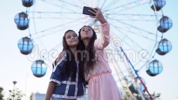 两个年轻女孩站在摩天轮的背景上用智能手机自拍慢动作视频的预览图