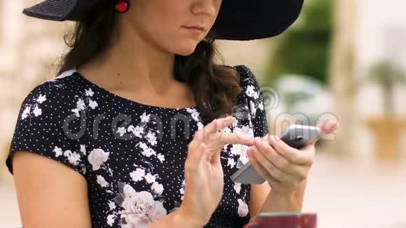 漂亮的女士戴着帽子坐在街头咖啡馆滚动智能手机应用程序视频的预览图