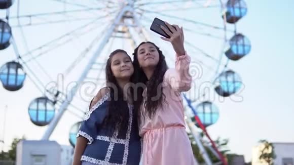 两个女孩站在摩天轮的背景上用智能手机自拍慢动作视频的预览图