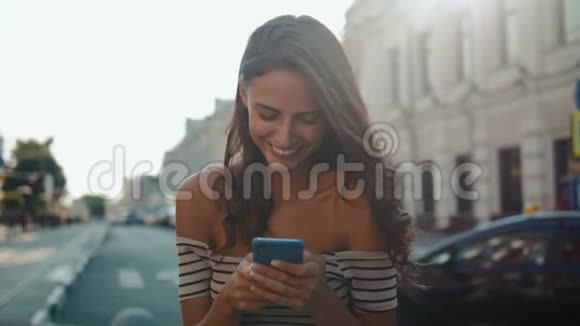 开朗的年轻女人穿着裙子在阳光明媚的城市街道上使用她的手机喝着纸杯里的咖啡视频的预览图