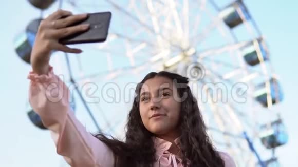 一个留着长发的女孩在摩天轮附近站着用手机自拍慢动作肖像视频的预览图