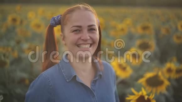 带着红色尾巴的年轻女人站在田野里向日葵在夕阳下微笑特写肖像视频的预览图