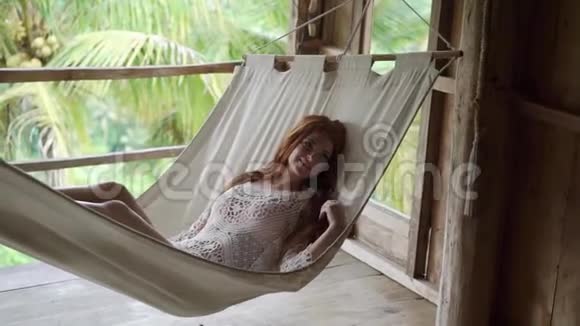 一个穿着透明浅色连衣裙躺在吊床上的年轻女孩红发美女度假视频的预览图