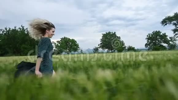 年轻快乐的金发女孩在雨天的背景下晚上跑在绿色的麦田上侧视图视频的预览图
