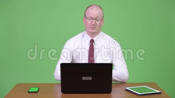 成熟的秃头商人在工作时使用笔记本电脑和电话靠木桌视频的预览图
