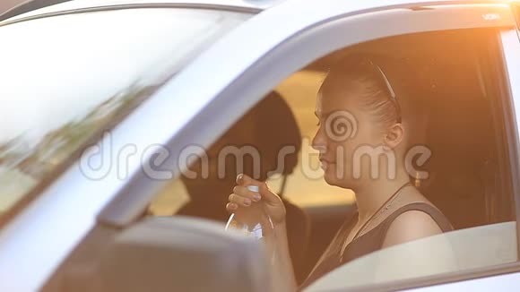 年轻的美女戴着墨镜戴着红发坐在汽车的车轮后面喝着瓶子里的水视频的预览图