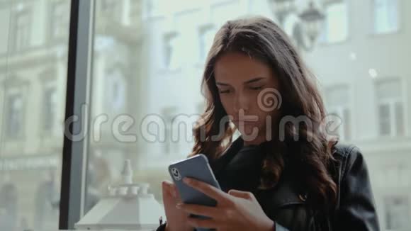 年轻微笑的商务女性坐在桌边的咖啡馆里把手靠在椅子上拿着智能手机在桌上视频的预览图