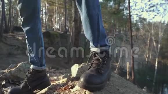 穿着靴子和蓝色牛仔裤的男腿站在悬崖边的岩石上山区靴子的绿景视频的预览图