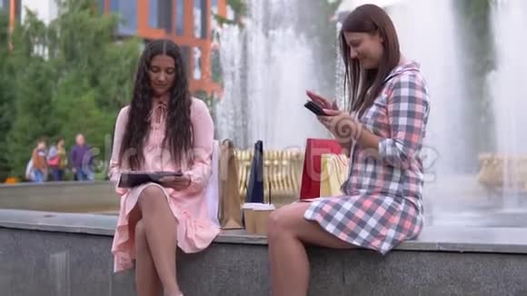 两个购物后的年轻女孩正坐在公园的喷泉附近慢动作高清视频的预览图
