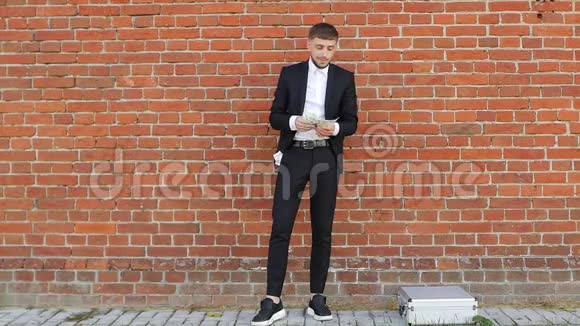 一个穿着黑色西装的年轻人在一堵墙的背景下数钱把自己的一半放在口袋里视频的预览图