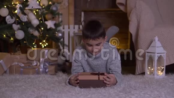 一个小男孩打开一个带礼物的盒子高兴地躺在圣诞树附近的地板上高清高清视频的预览图