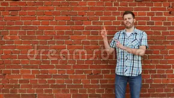 穿着衬衫和牛仔裤的时髦留胡子的人指出要复制空间砖墙背景视频的预览图