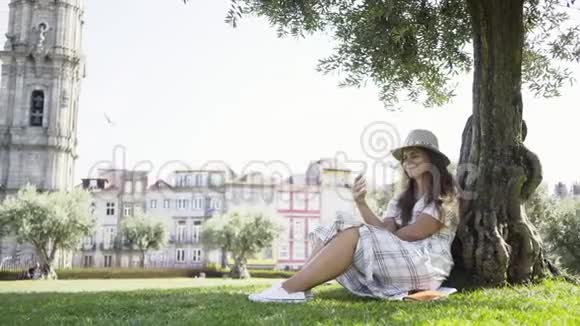 穿着智能手机的女人坐在公园的树下视频的预览图