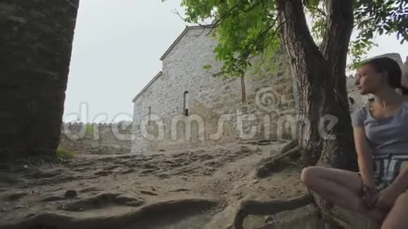 在古代防御堡垒的领土上女孩坐在树根上用石头砌成视频的预览图