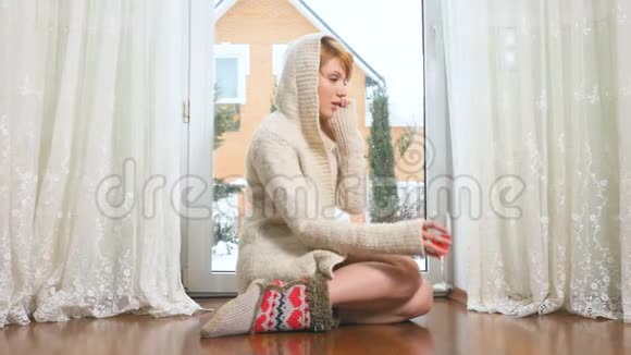 穿着毛衣的美女坐在靠近窗户的地板上打电话喝酒视频的预览图