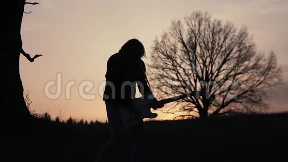 日落时分人们在靠近树的田野里弹着电吉他唱歌剪影视频的预览图