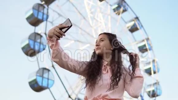 一个穿着粉红色连衣裙的长发女孩用站在摩天轮附近的智能手机自拍4K视频的预览图