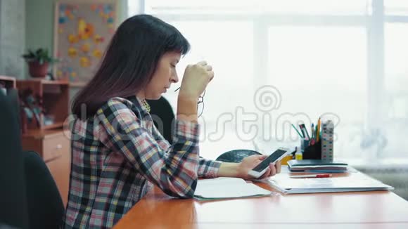 年轻的女老师坐在桌子旁处理文件和智能手机视频的预览图