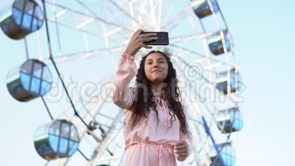 一个留着长发的女孩在摩天轮附近站着用手机自拍慢动作视频的预览图