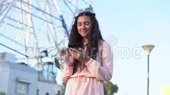 一个留着深色长发的女孩用智能手机站在大摩天轮和游乐园附近慢动作视频的预览图