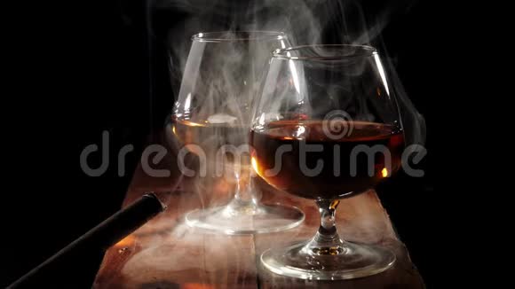 豪华白兰地两个带有金色干邑的玻璃杯和一支带有烟的雪茄放在黑色背景的棕色木桌上抽烟视频的预览图