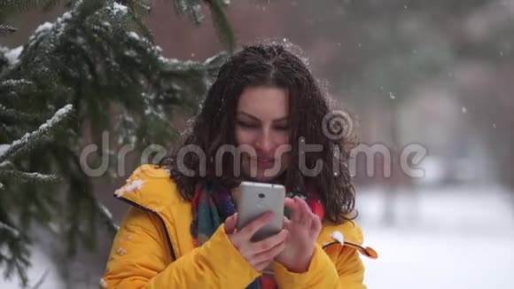 一个卷曲的黑发女孩在一场雪中带着智能手机在公园里散步的亲密肖像在雪的背景下视频的预览图
