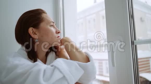 一个穿着白色外套的沮丧的女人坐在窗台上向外看着街道一个悲伤的女人坐在视频的预览图