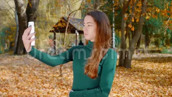 年轻漂亮的黑发女人在秋天的黄树背景下在智能手机上自拍视频的预览图