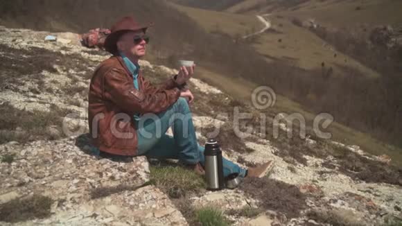 山上坐着一个男人戴着牛仔帽皮夹克蓝色牛仔裤和眼镜一个人喝热水瓶里的茶视频的预览图