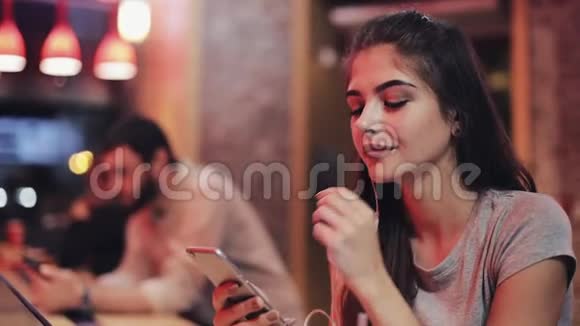 漂亮的年轻女人坐在酒吧后面用智能手机听音乐微笑喝鸡尾酒她是视频的预览图