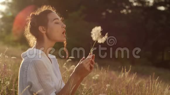 美丽的年轻女子在夏日夕阳下的麦田里吹蒲公英美丽与夏日概念视频的预览图