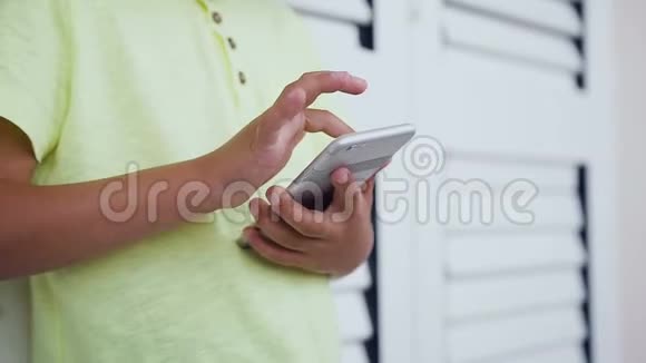 一个身高5到6岁在智能手机上玩网络游戏的男孩站在白色窗户附近视频的预览图