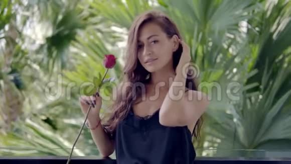 可爱的年轻女士穿着黑色睡衣在异国情调酒店的阳台上闻着红玫瑰视频的预览图
