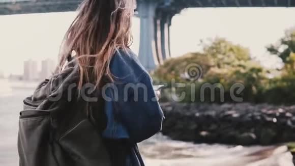 年轻女游客的侧视与背包行走在阳光明媚的布鲁克林公园河使用智能手机慢动作视频的预览图