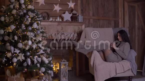 圣诞节一个女孩坐在圣诞树旁的椅子上喝茶视频的预览图