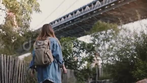 低角度拍摄的年轻旅行者女孩与背包下行走美丽的布鲁克林大桥公园树木慢动作视频的预览图