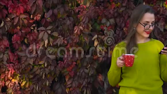戴眼镜的女人在户外喝茶或咖啡在温暖的秋日使用智能手机咖啡休息时间视频的预览图