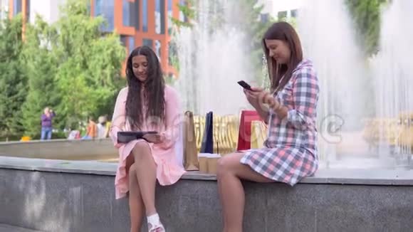 在阳光明媚的天气里两个购物后的漂亮女孩正坐在喷泉附近的公园里用手机和平板电脑视频的预览图
