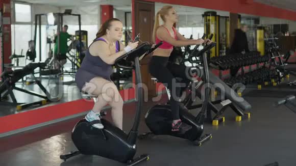 超重女士缓慢地骑着运动自行车在智能手机上滚动视频的预览图