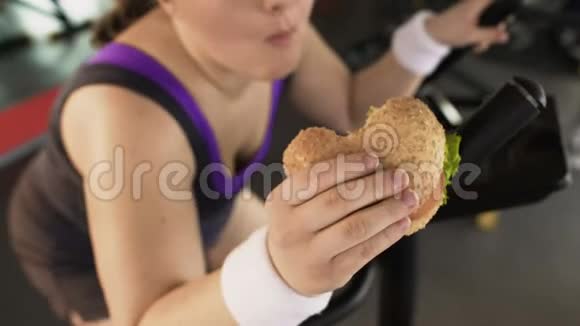 胖女人骑着固定的自行车在健身房吃美味的汉堡不健康的饮食视频的预览图