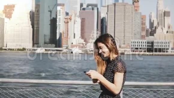 美丽的年轻白种人女人长发沿着纽约天际线全景在阳光明媚的一天使用智能手机视频的预览图